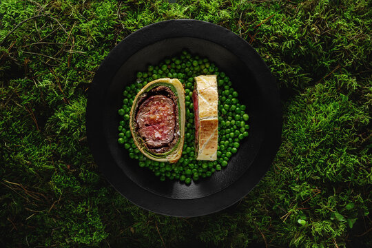 Homemade Christmas Beef Wellington as advent creation served in black plate with green pees on forrest moss  background. Food plating and aesthetic concept