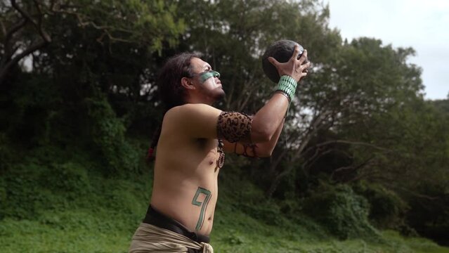 Mayan male warrior in traditional body costumes and face paint engaging in the ancient Mesoamerican ballgame with a dark ball in a grass near ancient ruins in Mexico
