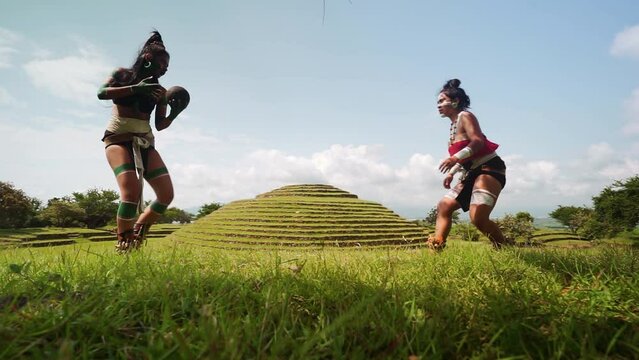 Mayan female warriors in traditional body costumes and face paint engaging in the ancient Mesoamerican ballgame with a dark ball in a grass near ancient ruins in Mexico