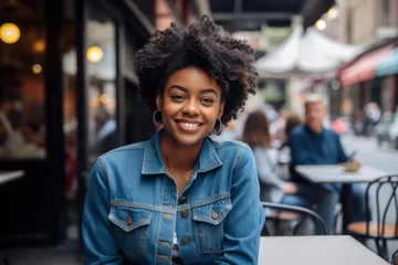 Tuinposter Portrait of a glad afro-american woman in her 20s sporting a versatile denim shirt against a bustling city cafe. AI Generation © Markus Schröder