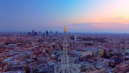 Aerial view of the statue of the Madonna on the central spire of the Duomo and the city's cathedral at dawn. Roofs of houses and skyscrapers. Sun over the horizon. duomo square. Italy Milan 16.11.2023