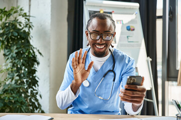 joyous african american doctor with glasses waving at mobile phone camera and smiling happily