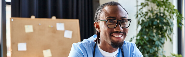 joyful african american doctor with glasses looking at camera during online consultation, banner