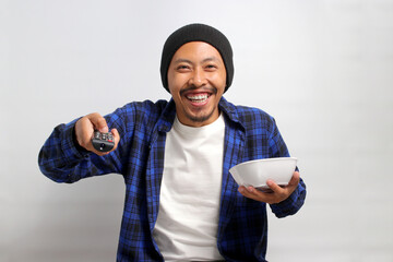 An enthusiastic young Asian man, dressed in a beanie hat and casual shirt, increases the TV volume...