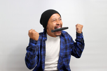 Funny young Asian man, dressed in a beanie hat and casual shirt, bites the TV remote in frustration...