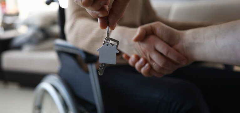 Close-up of persons hand giving keys from new apartment to disabled female. Woman sitting in wheelchair. Moving day into apartment. Handicapped and real estate concept