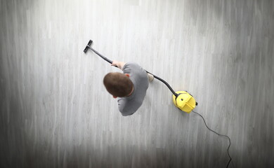 Top view of adult male cleaning in room with compact yellow vacuum cleaner. Grey laminate floor in...