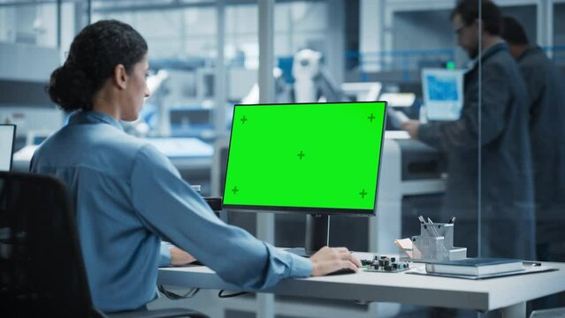 Hispanic Female Robotics Engineer Using Desktop Computer with Green Screen Chromakey Mock Up Display in Factory Office. Technological Research and Development Facility with Autonomous Assembly Line.