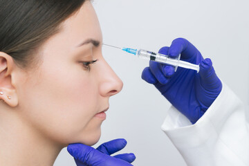 A cosmetologist in blue gloves gives a pretty young woman  botox injections into her forehead with a syringe to correct her wrinkles between her eyebrows. Aesthetic cosmetology. Face in profile.