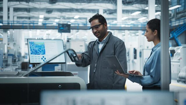 Indian Male Industrial Engineer And Hispanic Female Supervisor Using Laptop And Talking At Electronics Factory. Man Using Soldering Jet Printer, Explaining Process Behind Production Of Motherboards.