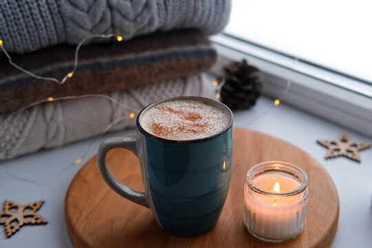 Winter windowsill still life. Blue ceramic cup of hot coffee on window sill. Christmas decorations on the background. Cozy home picture. Warm woolen knitted sweaters, Burn Candle, Cookies. Stock photo