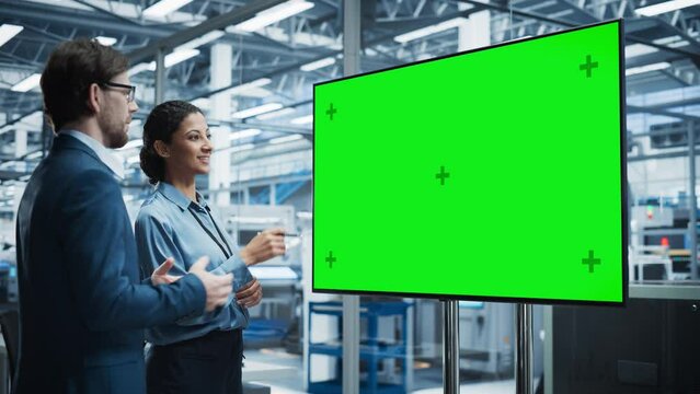 Meeting In Office Of Autonomous Factory With Robotic Arms: Hispanic Female Chief Engineer Talking with Caucasian Male Business Partner in a Conference Room with TV with Green Screen Mock Up Display.
