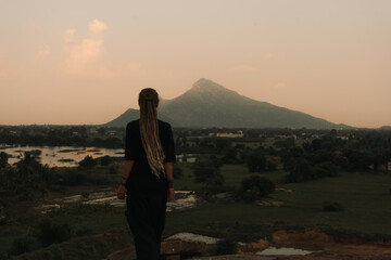 Young woman traveler wearing dreadlocks standing in tropical landscape in front of sacred mountain...