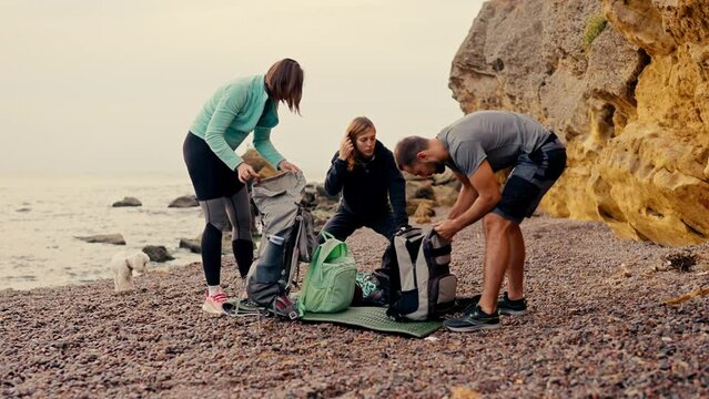 A group of rock climbers unpack their backpacks and prepare to climb the yellow rocks near the sea on a rocky shore in summer. A blonde girl in a black sports uniform, as well as a girl with a bob