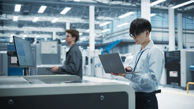 Asian Male Engineer Using Laptop Computer To Control Soldering Jet Printer On Electronics Factory. Man Working On Production Of Innovative Printed Circuit Boards For Artificial Intelligence Systems.