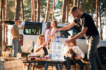 Sitting by the table with food, against car. Group of friends are together in the forest