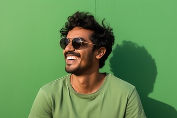 Portrait of a smiling indian man in his 20s wearing a trendy sunglasses against a soft green background. AI Generation