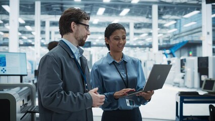 Caucasian Man and Hispanic Female Having a Conversation at an Industrial Facility. Advanced Factory Employees Working Together, Using Laptop Computer for Supervising Assembly Process Of New Devices.