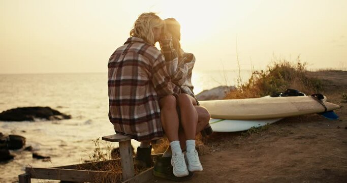 Happy couple a blond man in a checkered shirt sits on a wooden bench on his legs sits a blonde girl in a checkered shirt and a hat they hug and kiss near their surfboards on strawberries near the sea