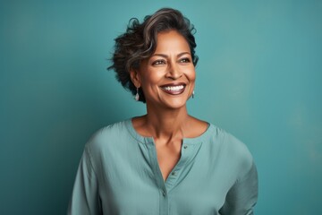 Fototapeta premium Portrait of a smiling indian woman in her 50s sporting a long-sleeved thermal undershirt against a soft teal background. AI Generation