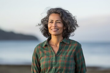 Portrait of a satisfied indian woman in her 50s wearing a comfy flannel shirt against a serene seaside background. AI Generation