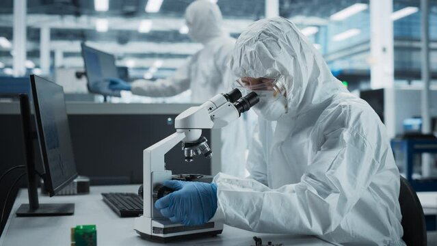 Two Scientists In Sterile Suites At Electronics Factory: Engineer Using Microscope And Desktop Computer To Inspect Microchip While His Colleague Using Soldering Jet Printer To Print Modern Motherboard