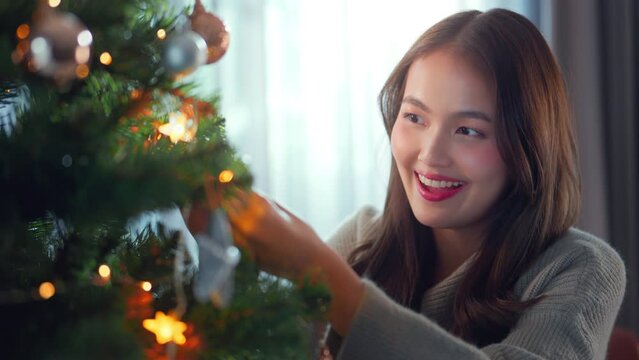 Happy Young asian woman wearing Santa hat hangs up golden ball on branch of christmas tree, house decoration, preparing to celebrate Christmas and New Year at home.