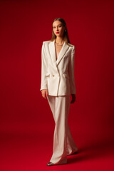 Fashionable confident woman wearing trendy white suit double breasted blazer, classic trousers, metallic color shoes, posing on red background