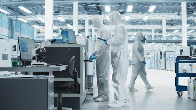 Two Specialists in Sterile Suits Walking In Electronics Factory Research Center. Scientist With Tablet And Engineer Approaching Soldering Jet Printer And Starting Production Process Of Circuit Boards.