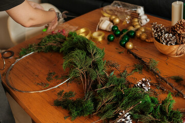 A young woman in an apron assembling a Christmas wreath from fir branches. Decorations for the New Year are scattered on the table: pine cones, Christmas tree balls and a star.