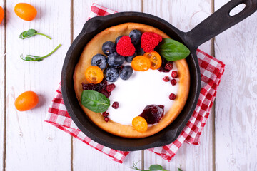 Dutch baby pancake served with custard, whipped cream and berries in cast iron pan on white wooden table. Top view