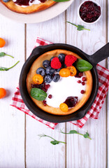 Dutch baby pancake served with custard, whipped cream and berries in cast iron pan on white wooden table. Top view