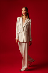 Fashionable confident woman wearing trendy white suit double breasted blazer, classic trousers, metallic color shoes, posing on red background