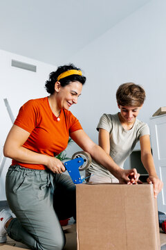 Focused mother in red top packaging box with tape dispenser with son