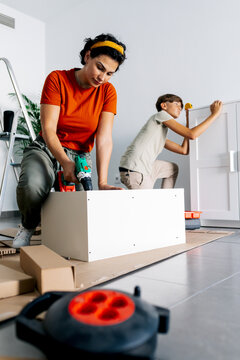 Serious single parent with son assembling cabinet with drill machine during relocation