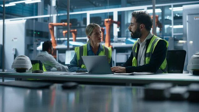 Autonomous Factory Office: Caucasian Female Quality Control Technician Talks to Hispanic Male Industrial Engineer Working on Laptop Computer. Automated Robot Arm Assembly Line Manufacturing Machinery.