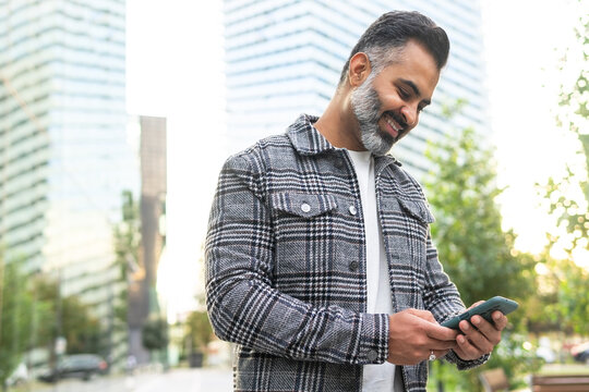 Bearded Indian businessman using cellphone and smiling while standing outside