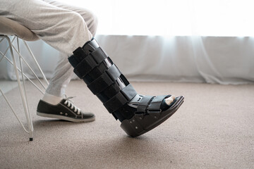 Foot orthosis concept. Close-up of man leg with ankle orthosis, black splint, walker boot on the sofa, profile view. - obrazy, fototapety, plakaty