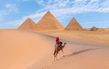 Camels in Giza Pyramid Complex - A woman in a red turban riding a camel across the thin sand dunes - Cairo, Egypt