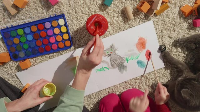 Mothers and childs hands in process of painting their family pet, gray cat lies near, top view, furry white carpet background, wooden toy blocks around, first arts with parents help. HQ 4k footage