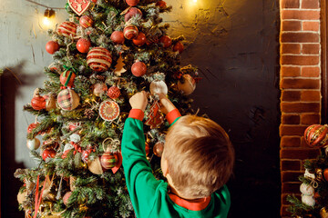 unrecognizable Child hanging a ball on illuminated Christmas tree.glowing lights. Boy in green pajamas decorating Christmas tree.Christmas good mood. Lifestyle, family and holiday.