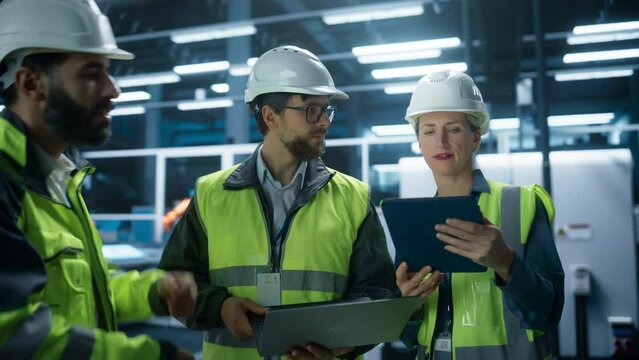 Meeting Of Diverse Engineers In Autonomous Factory Hall: Male And Female Technicians Using Tablet, Laptop And Discussing Industrial Machinery Production On Automated Assembly Line With Robotic Arms.
