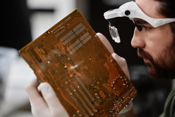 Side view closeup of repairman inspecting circuit board with magnifying glasses in electronics...