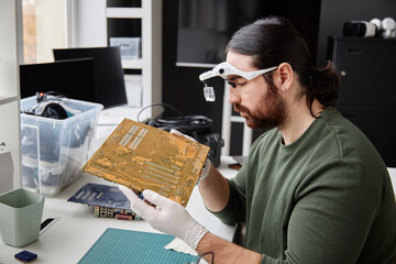 Side view portrait of bearded repairman inspecting circuit board with magnifying glasses in...