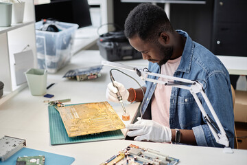 Side view portrait of African American man fixing circuit board with soldering iron in computer...