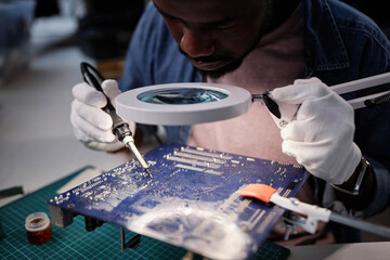 Close up of African American man inspecting circuit board with magnifying glass in computer repair workshop, copy space - Powered by Adobe
