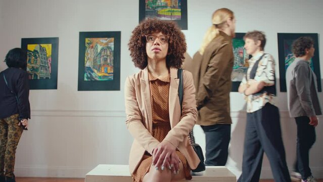 Time lapse of female visitor sitting in exhibition hall of art gallery, looking at paintings with admiration while people walking behind