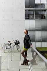 Stylish and pretty young woman in black jacket and silk dress standing near bicycle in Berlin