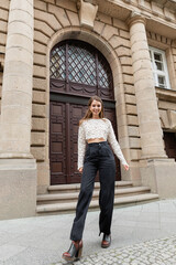 Fototapeta na wymiar cheerful young woman in lace top and high waist pants looking at camera on urban street in Berlin