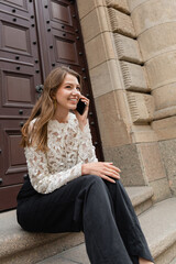 Fototapeta premium Cheerful woman talking on smartphone while sitting on stairs near entrance of building in Berlin